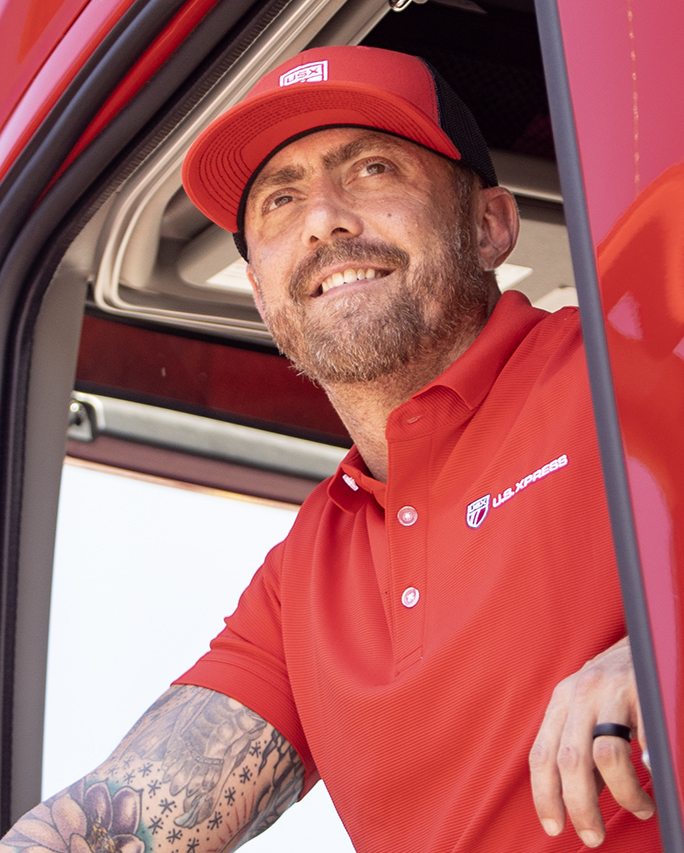 US Xpress driver with red shirt and hat on looks out of his truck.
